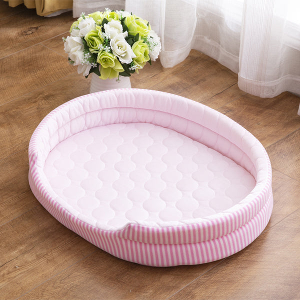 Pink and white striped Cove Cooling Cat Bed with a quilted cooling pad, on a wood floor by a window