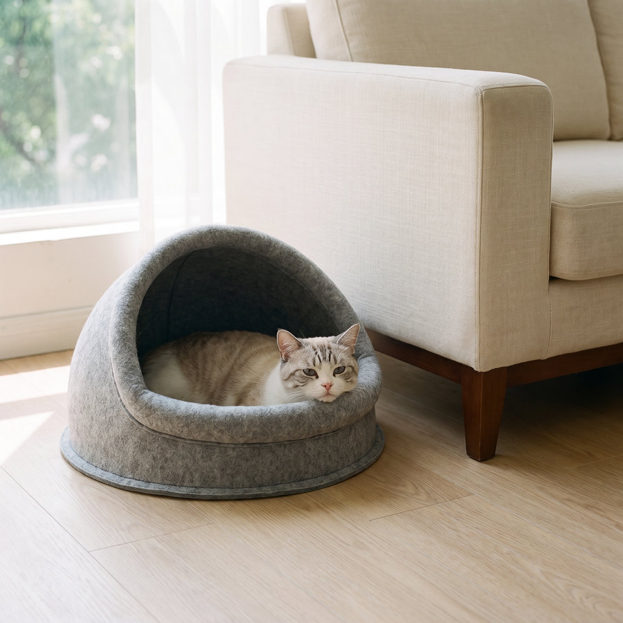 A cat rests in the FELVO Fold semi-enclosed grey felt cat cave beside a cream sofa